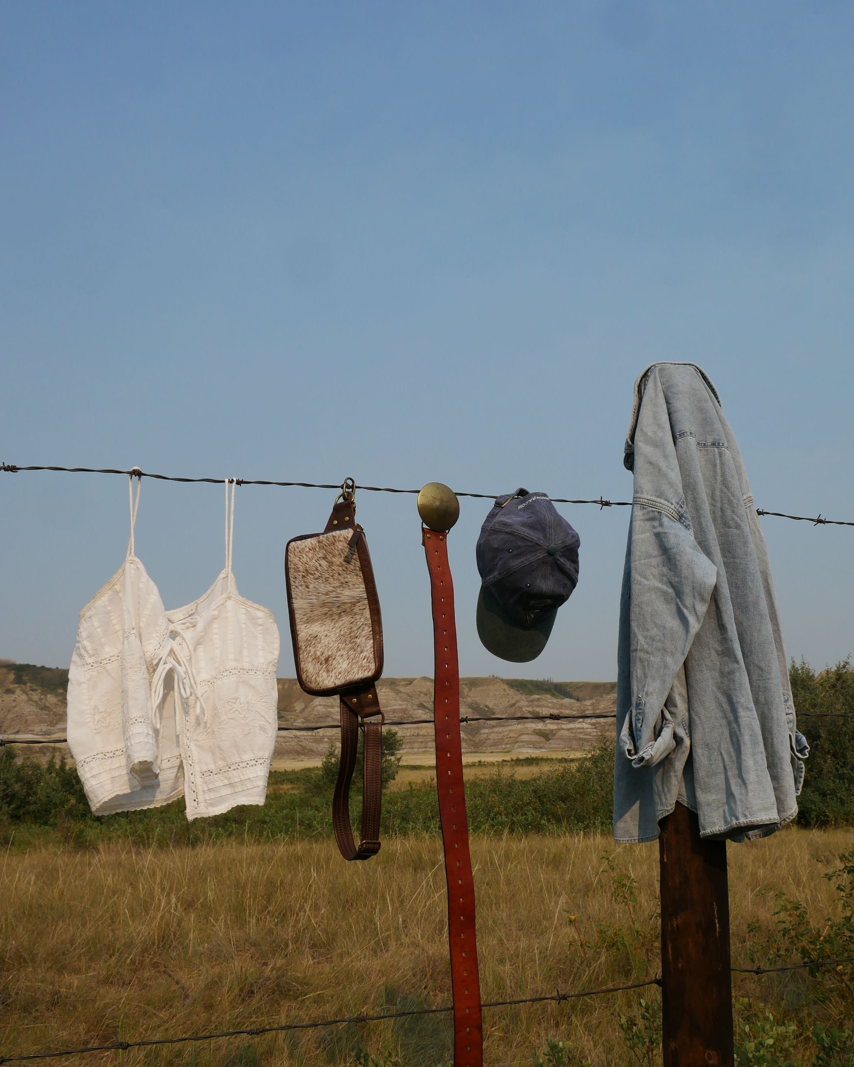 A horizontal outdoor scene showing several pieces of clothing and a small bag hanging from a wire fence in a grassy field under a blue sky. Curated style bundle for Just Dandi client.