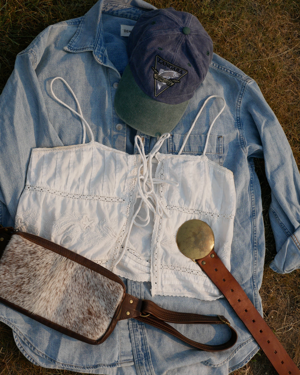 A styled flat-lay of western-inspired accessories on a light blue denim shirt laid on dry grass, including a white lace-up corset-style vest, a brown leather belt with a large round metal buckle, a brown-and-white cow hide shoulder bag, and a blue baseball cap. Carefully curated as a style bundle