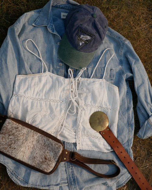 A styled flat-lay of western-inspired accessories on a light blue denim shirt laid on dry grass, including a white lace-up corset-style vest, a brown leather belt with a large round metal buckle, a brown-and-white cow hide shoulder bag, and a blue baseball cap. Carefully curated as a style bundle