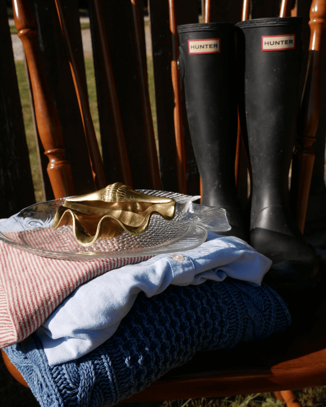 A pair of black Hunter rain boots standing on a wooden chair next to a stack of folded knit sweaters and a gold-toned decorative object on a glass plate. Curated for a client to match their Pinterest board, curated thrift style bundle.