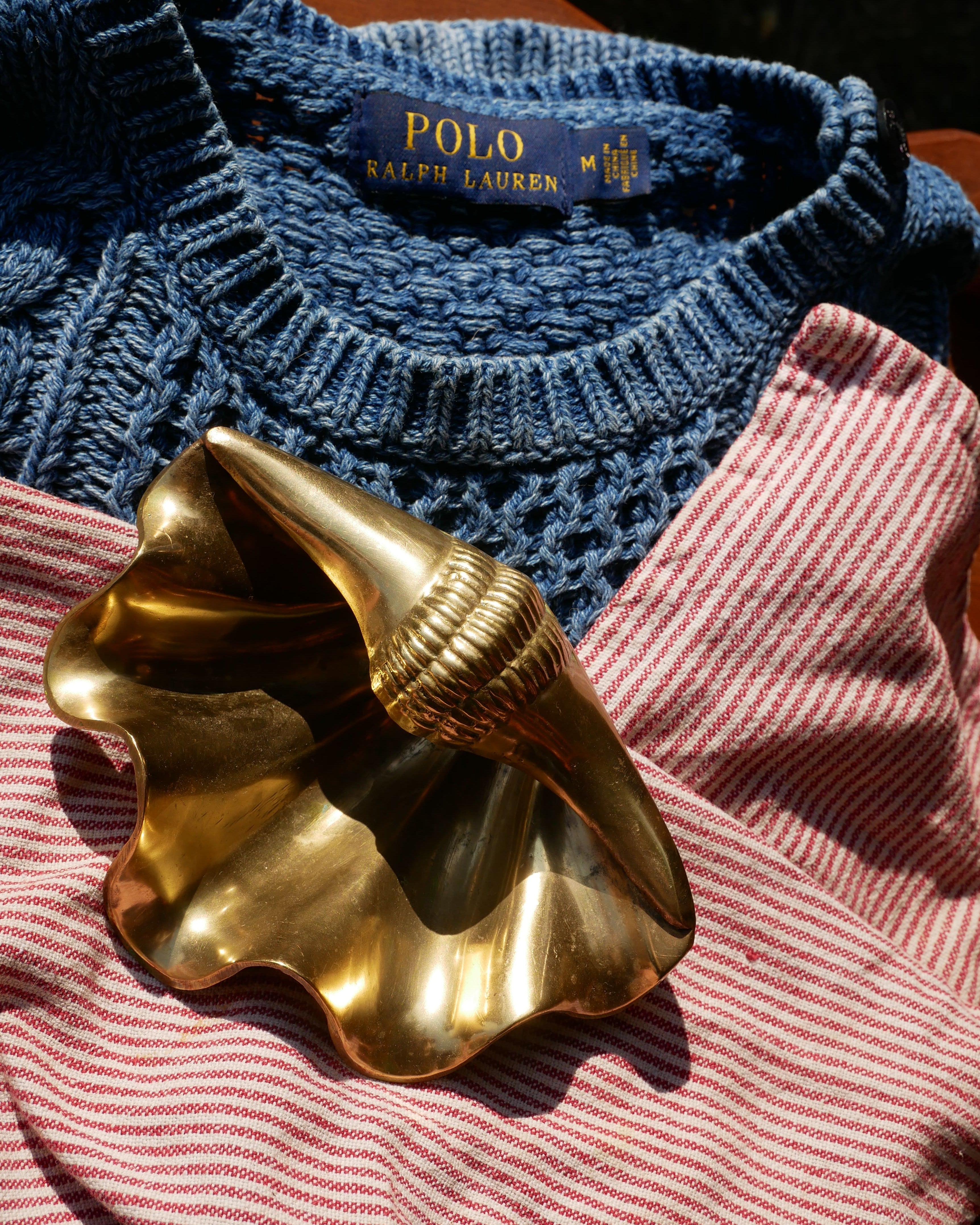 A close-up of a blue knit sweater with a visible Polo Ralph Lauren neck label, laid over a pink and white striped fabric, with a shiny gold shell-shaped object resting on top. Curated style bundle from Just Dandi Curation, thrifted bundle for a clients personal style.