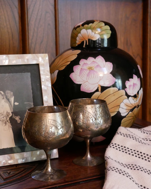 A styled tabletop arrangement featuring two engraved silver goblets, a black lacquered floral jar, a framed black-and-white wedding photograph, and a cream knit blanket with black dots, set against a dark wood paneled background. For a curated thrifted style bundle, tailored specifically to clients style.
