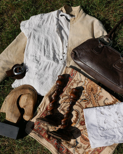 A styled flat-lay of vintage accessories and textiles on green grass, including a brown leather handbag, a patterned rug, a wooden spoon, a white lace-trimmed dress, and a small camera. Ralph Lauren inspired style bundle, curated thifted style bundle.